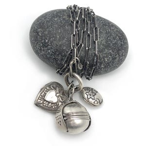 Peut inclure: Un collier en argent avec une chaîne et plusieurs breloques, dont un médaillon en forme de cœur, un médaillon rond et un charm floral. Le collier est posé sur une pierre grise sur fond blanc.