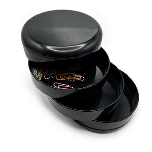 May include: A black, round, tiered storage container with three open trays and a lid. The trays are filled with colorful paper clips. The container is made of a glossy material and sits on a white background.
