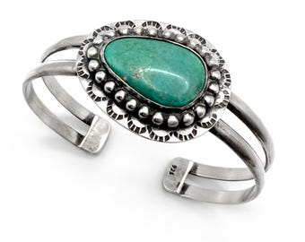 May include: A silver cuff bracelet with a large oval turquoise stone at the center. The stone is surrounded by a beaded and scalloped silver setting. The bracelet has a split design with two parallel bands on each side.