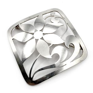 Può includere: Una spilla quadrata in argento con un design intagliato raffigurante un fiore centrale a sei petali e steli curvi a forma di foglia. Il metallo presenta una finitura liscia e riflettente, con angoli arrotondati e spazi vuoti definiti.