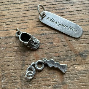 May include: Three silver charms on a wooden surface. One charm is a small shoe, another is a rectangular tag with the words "Follow your bliss," and the third charm reads "LOVE IS BLIND."