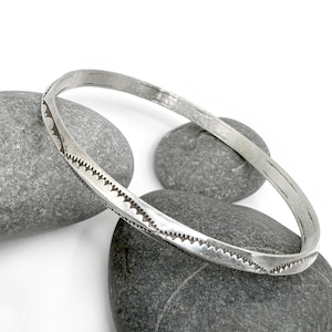 May include: A silver-toned bangle bracelet with a textured surface and a decorative edge. The bracelet is resting on gray stones against a white background. The bracelet's design features a series of small, repeating shapes along its edge.