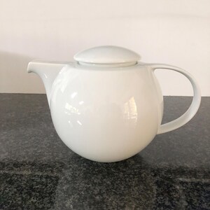 May include: A white ceramic teapot with a rounded body, a curved handle, and a spout. The teapot has a matching lid and sits on a dark speckled surface. The design is minimalist and modern.
