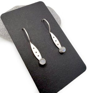 May include: A pair of silver earrings with a curved hook design. Each earring features an elongated, oval-shaped body with three small, evenly spaced holes and a small, round, light blue gemstone at the bottom. The earrings are displayed on a black surface.