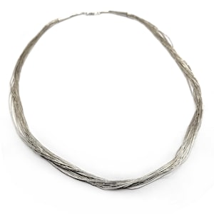 Puede incluir: Un collar plateado compuesto por numerosos hilos finos, formando un diseño circular y en capas. El collar se presenta sobre un fondo blanco, resaltando su delicada estructura y brillo metálico. El cierre es visible en la parte posterior.