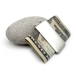 Könnte beinhalten: Ein silberner Geldklammer hält zwei US-Dollar-Scheine, neben einem glatten, grauen Stein. Die Scheine sind teilweise sichtbar, mit dem Text "SERIES 2017 A" und "Steven T. Mnuchin, Secretary of the Treasury".