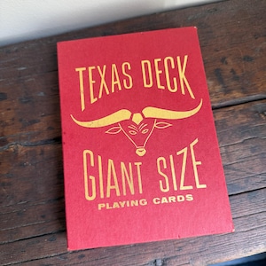 May include: A red playing card box with gold lettering that reads "TEXAS DECK" above a bull's head graphic and "GIANT SIZE PLAYING CARDS". The box is on a wooden surface.