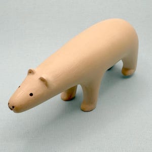 Könnte beinhalten: Eine glatte, hellbeige Holzfigur in Form eines Eisbären in Laufhaltung. Der Bär hat einen langen, abgerundeten Körper, vier kräftige Beine und einen einfachen Kopf mit zwei kleinen Ohren sowie zwei dunklen, kreisförmigen Punkten als Augen.