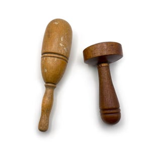 Könnte beinhalten: Zwei Küchenwerkzeuge aus Holz auf weißem Hintergrund. Eines ist ein hellfarbiger, länglicher Stößel mit Griff. Das andere ist ein dunkel gefärbtes, pilzförmiges Werkzeug mit Griff. Beide Werkzeuge scheinen Vintage zu sein.