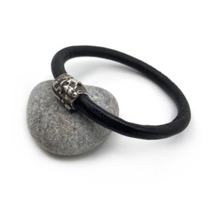 Op de afbeelding: Een zwart leren armband met een zilverkleurige sluiting. De armband ligt op een grijze steen tegen een witte achtergrond. De sluiting heeft een textuur.