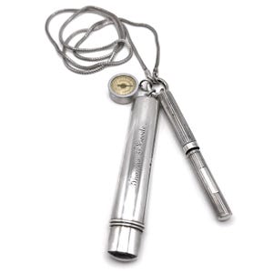 May include: A silver pendant necklace featuring a compass, a cylindrical case with the engraved text "Thomas F. Goode", and a pen. The compass has a cream-colored face. The pen has vertical lines. The necklace chain is a silver color.
