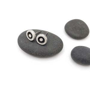 May include: A pair of circular stud earrings featuring a silver outer ring and a dark recessed center with a small silver dot in the middle. The earrings have a post back design and a polished metallic finish, showcasing a simple geometric pattern.