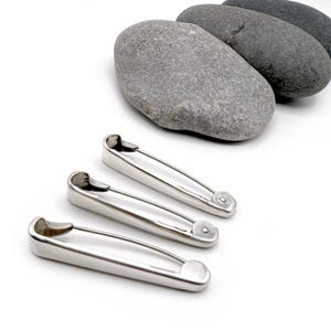 May include: Three shiny silver safety pins are arranged in the foreground, with three gray stones in the background. The safety pins have a classic design with a curved wire and a secure clasp.