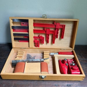May include: A wooden tool box containing various red plastic clamps, a metal saw blade, two cylindrical sanding blocks, and two red metal hand tools. The box features a central wooden block with the embossed text "model power" on its surface.