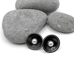 Könnte beinhalten: Ein Paar dunkel versilberte Ohrstecker mit Perlen in der Mitte. Die Ohrringe haben ein ausgestelltes, schalenförmiges Design. Im Hintergrund befinden sich mehrere graue Steine.