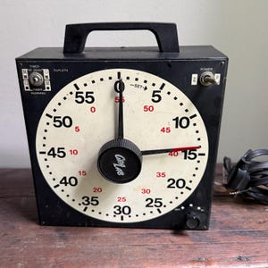 May include: A black GraLab darkroom timer with a square housing and a top handle. The circular face features a white background with large black numbers for seconds and smaller red numbers for minutes. It includes a central control knob, a power toggle switch, and a black electrical cord.