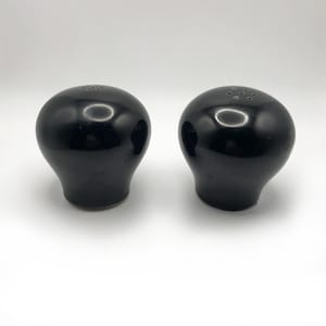 Könnte beinhalten: Zwei schwarze Salz- und Pfefferstreuer aus Keramik. Jeder Streuer hat eine abgerundete Oberseite mit kleinen Löchern und einen bauchigen Boden. Die Streuer sind glänzend und reflektieren das Licht, vor einem weißen Hintergrund. Das Design ist schlicht und modern.