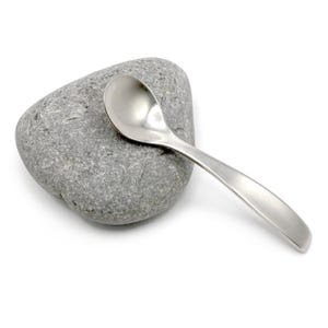 May include: A small, polished silver-toned metal spoon resting on top of a smooth, grey, irregularly shaped river stone. The spoon features a rounded bowl and a curved handle, contrasting with the textured, matte surface of the natural stone.