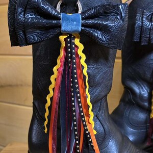 May include: A black leather cowboy boot featuring an embossed floral pattern, adorned with a large black bow attached to a silver ring. A cluster of dangling ribbons in yellow, orange, red, and black with white polka dots hangs from the center.