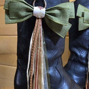 May include: A black leather cowboy boot adorned with a decorative accessory featuring a large olive green fabric bow. The center of the bow has a white ribbon with small leaf patterns, and a long tassel made of thin, multicolored ribbons hangs down.