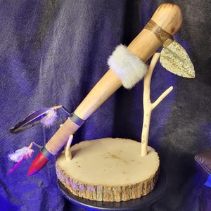 May include: A wooden ceremonial tomahawk with a brown leather wrap and a stone blade. The tomahawk is mounted on a wooden stand with a branch and fur accents.
