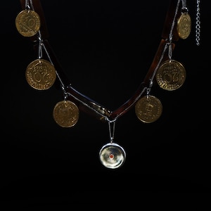Peut inclure: Un collier unique avec un design géométrique marron foncé, orné de pièces de monnaie dorées et de chaînes argentées. Un pendentif argenté avec une petite pierre rouge est suspendu au centre. Le fond est noir uni.