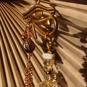 Könnte beinhalten: Ein goldener Anhänger mit einem ägyptischen Horus-Augen-Motiv, der an einer Kette mit verschiedenen Perlen hängt, darunter eine braune Muschelperle, eine weiße Perle und klare Quarzperlen.