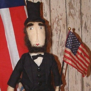 Puede incluir: Muñeca artesanal de Abraham Lincoln, vestida con un traje negro y sombrero de copa, sosteniendo una pequeña bandera estadounidense. La muñeca tiene una cara de tela con barba y está frente a una bandera estadounidense.