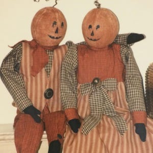 May include: Two fabric dolls with round orange pumpkin heads featuring hand-painted black facial features and wire stems. The dolls wear outfits made of brown, orange, and tan striped and checkered fabric, with black fabric hands and feet.