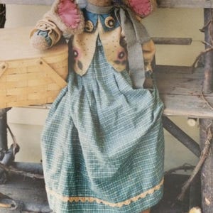 May include: A handmade Easter bunny doll with long, floppy ears and pink inner ears. The bunny wears a blue and green plaid dress, a vest with floral accents, and tan pants. A small basket sits nearby, suggesting a springtime theme.