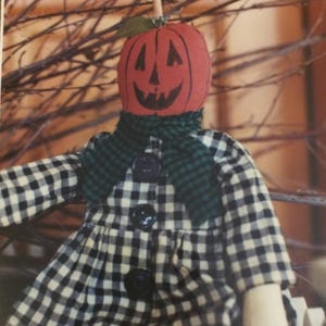 May include: A handmade Halloween decoration featuring a pumpkin head with a carved face. The figure wears a black and white checkered dress with large black buttons and a green plaid scarf. The background includes bare branches.