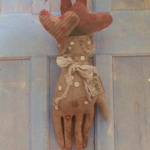 May include: A decorative fabric hand with two red heart-shaped accents. The hand is made of tan burlap-like material, adorned with white buttons, lace, and a small tag. A white ribbon is tied around the wrist area. The background is a light blue wooden surface.