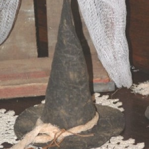 May include: A black, pointed witch hat with a textured, weathered appearance. The hat features a wide brim wrapped with a thick, light-colored, frayed jute or twine band that is tied in a knot at the front, adding a rustic and worn aesthetic.