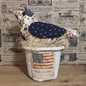 May include: A patriotic fabric bird figurine with a star and stripe pattern, perched in a white pot. The pot features an American flag design and the words "OLD GLORY". The bird sits on a bed of straw.