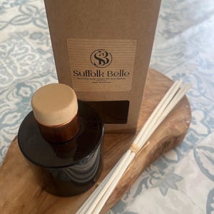 May include: A dark brown glass diffuser bottle with a wooden cap and copper trim, beside a kraft paper box and a bundle of diffuser reeds. The box has a label that reads "Suffolk Belle". The items are on a wooden board.