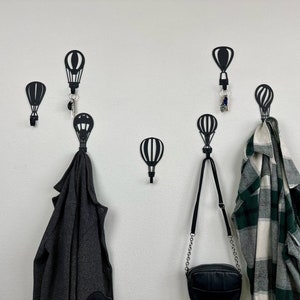 May include: Six black metal hot air balloon hooks mounted on a white wall. Two hooks have items hanging from them, a gray jacket and a green and white plaid shirt. A black purse with a silver chain strap hangs from a third hook.