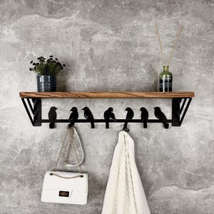 Op de afbeelding: Een wandplank met een houten bovenkant en zwarte metalen steunen. Daaronder een rij zwarte vogelvormige haken met een witte handtas en een handdoek. Een kleine plantenpot en een glazen fles met riet staan op de plank.