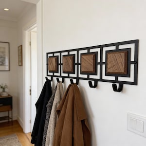 May include: A wall-mounted coat rack featuring a black metal rectangular frame with a geometric grid pattern. Four square wooden panels with a natural grain are centered within the grid, and five black metal hooks hang from the bottom edge.
