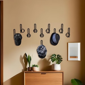 Könnte beinhalten: Eine Wandgarderobe mit schwarzen Metallhaken, an denen verschiedene Mützen hängen. Dazu gehören eine schwarze Baseballkappe, eine gemusterte Kappe und ein blau-schwarzer Fedora. Die Garderobe befindet sich über einem Holzschrank.