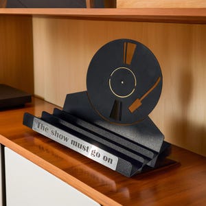 May include: Black and silver decorative record player sculpture with the text "The show must go on". The sculpture features a black record with gold and brown accents, and a silver base.