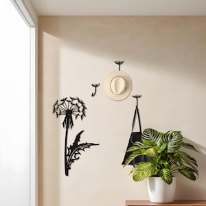 May include: Black metal dandelion wall art and hooks. A decorative dandelion silhouette is mounted on the wall, along with three dandelion-shaped hooks. A straw hat and a black tote bag hang from the hooks. A potted plant sits on a wooden surface.