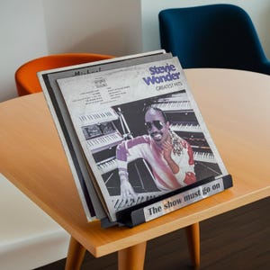 May include: A collection of vinyl records, including a Stevie Wonder "Greatest Hits" album, displayed in a black metal record holder. The holder is on a light wood table. The text "The show must go on" is visible on the holder.