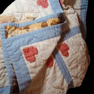 May include: A white quilt with a blue border and red embroidered hearts. The quilt is made of cotton and has a patchwork design. The quilt is folded in half and the edges are visible.