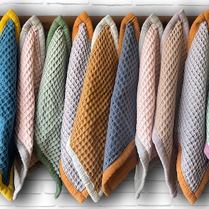 Peut inclure: Dix couvertures pour bébé en tissu gaufré, de différentes couleurs, suspendues à un support en bois. Les couleurs des couvertures sont le bleu sarcelle, le vert, le rose, l'orange et le gris. Chaque couverture est bordée d'une couleur contrastante.