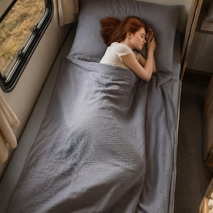 Puede incluir: Un juego de cama de muselina gris sólido que consta de una sábana encimera, una sábana bajera y una funda de almohada. La tela presenta una superficie arrugada y texturizada sobre un colchón. La ropa de cama es de un color gris neutro.