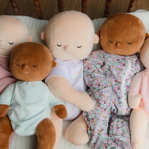 Puede incluir: Una colecci&oacute;n de mu&ntilde;ecos de peluche para beb&eacute;s con los ojos cerrados, en varios tonos de piel y atuendos. Los mu&ntilde;ecos visten un mono rosa, un body azul claro, una camiseta blanca y un saco de dormir con estampado floral.