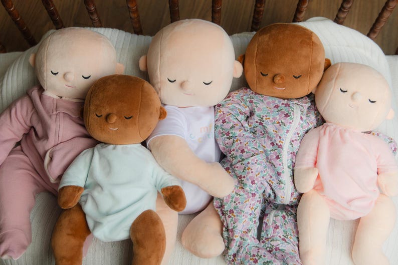 Puede incluir: Una colecci&oacute;n de mu&ntilde;ecos de peluche para beb&eacute;s con los ojos cerrados, en varios tonos de piel y atuendos. Los mu&ntilde;ecos visten un mono rosa, un body azul claro, una camiseta blanca y un saco de dormir con estampado floral.