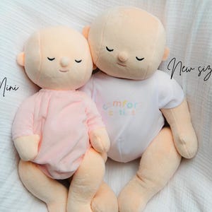 Puede incluir: Dos mu&ntilde;ecos de peluche, uno mini y otro de nuevo tama&ntilde;o, se muestran uno al lado del otro. El mu&ntilde;eco mini lleva un conjunto rosa, mientras que el m&aacute;s grande lleva un body blanco con el texto "comfort cuties". Ambos mu&ntilde;ecos tienen los ojos cerrados y piel color melocot&oacute;n.