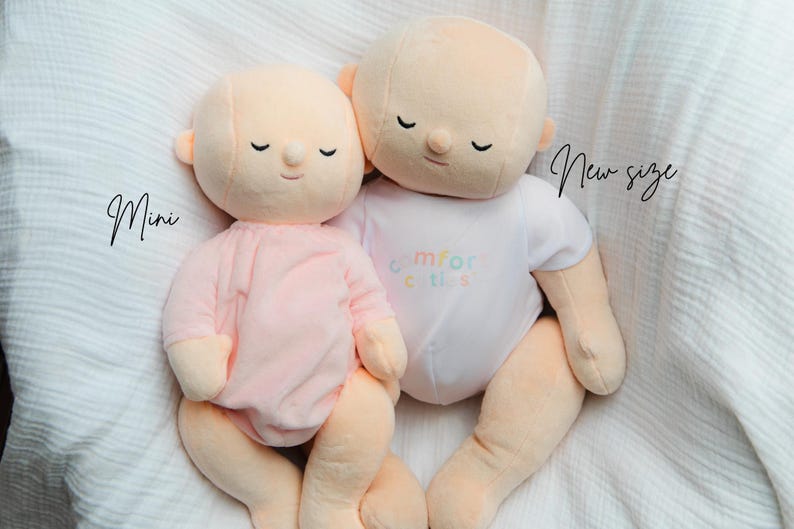 Puede incluir: Dos mu&ntilde;ecos de peluche, uno mini y otro de nuevo tama&ntilde;o, se muestran uno al lado del otro. El mu&ntilde;eco mini lleva un conjunto rosa, mientras que el m&aacute;s grande lleva un body blanco con el texto "comfort cuties". Ambos mu&ntilde;ecos tienen los ojos cerrados y piel color melocot&oacute;n.