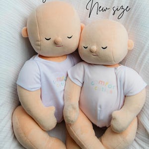 Puede incluir: Dos mu&ntilde;ecos de peluche con piel color melocot&oacute;n y ojos cerrados, que visten camisetas blancas con el texto "comfort cuties" en colores pastel. Los mu&ntilde;ecos est&aacute;n sentados sobre una superficie blanca y texturizada. Los textos "Original" y "New size" est&aacute;n escritos encima de los mu&ntilde;ecos.